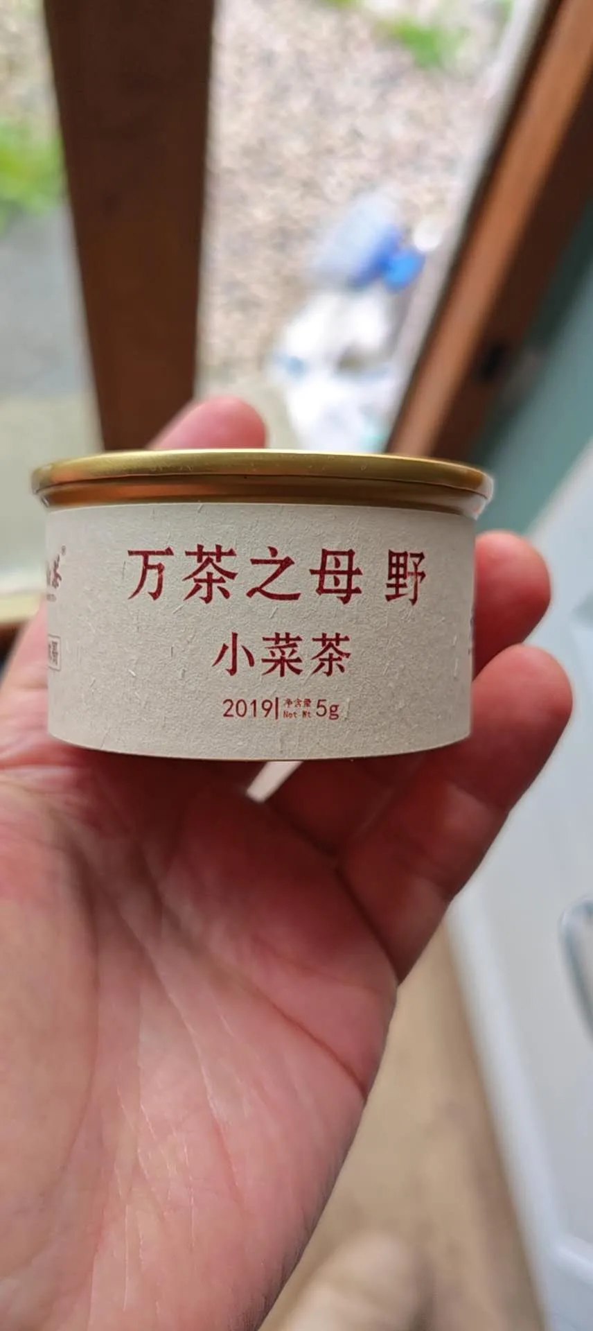 一只手托着一个小圆罐,金色金属盖,米白色纸标上印着红字「万茶之母 野」「小菜茶」,下面一行小字 2019 / 净含量 5g